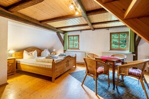 A wooden-framed room with a bed, wicker chairs, a table, and two windows with green curtains.