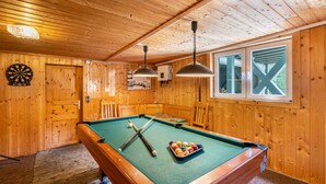 A pool table in a wooden room with a dartboard, a window with a view, and a ceiling light fixture.