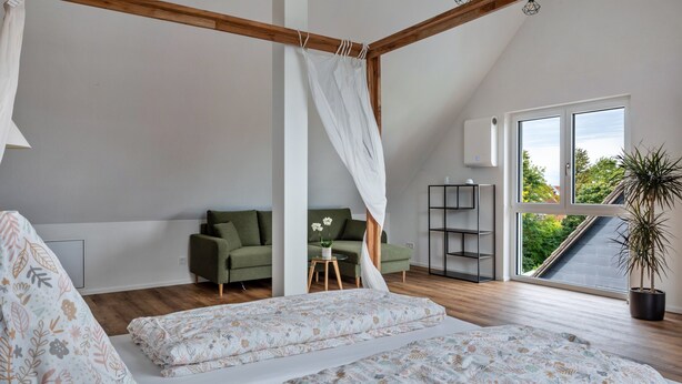 A bedroom with a canopy bed, a green sofa, a small table with flowers, a potted plant, and a view of greenery through a large window.