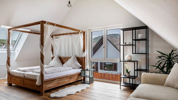 Ein Schlafzimmer mit einem hölzernen Himmelbett, einem Sofa, einem kleinen Beistelltisch, einer Topfpflanze und Blick auf ein Haus durch ein großes Fenster.