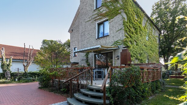 Ein Haus mit einer Steintreppe, die zu einer Holzdiele führt, umgeben von Grünpflanzen und einer gepflasterten Auffahrt.