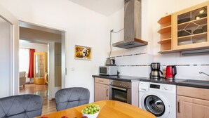 Eine kompakte Küche mit Mikrowelle, Toaster, Kaffeemaschine und Waschmaschine. Ein Esstisch mit Obstschale und einem Teller Äpfel.