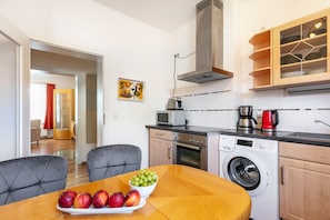 A compact kitchen with a microwave, toaster, coffee maker, and washing machine. There is a dining table with a bowl of fruit and a plate of apples.