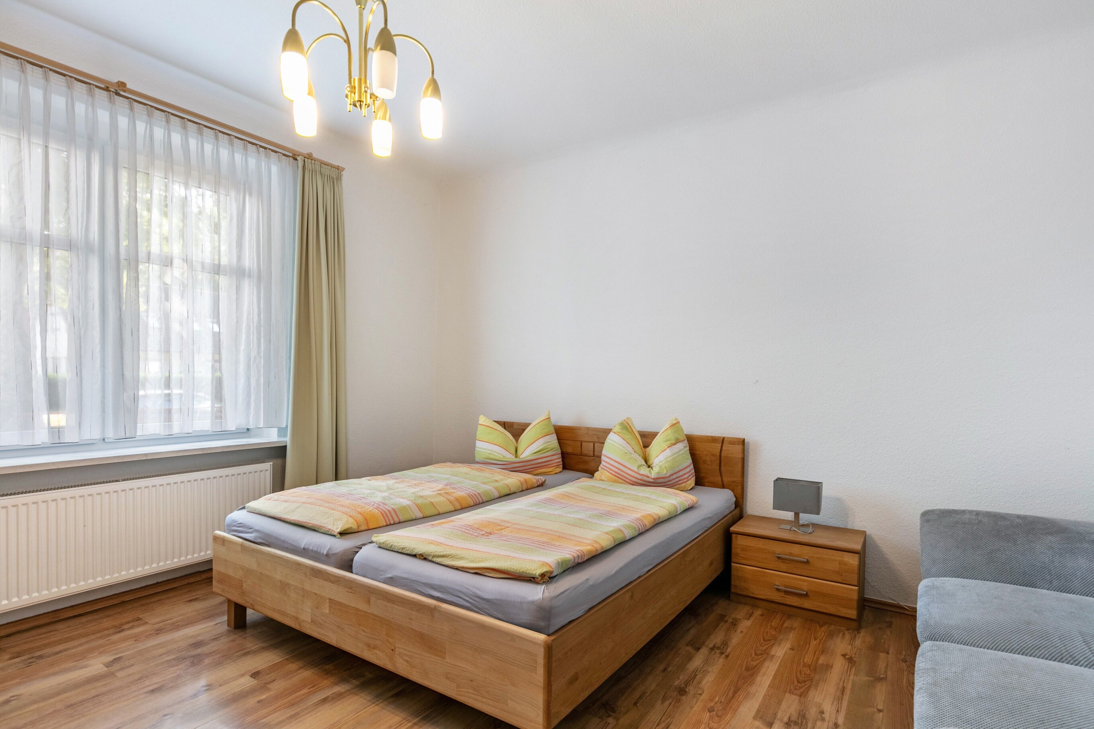 Una camera da letto con un letto di legno, un comodino, un divano grigio e una finestra con tende trasparenti.