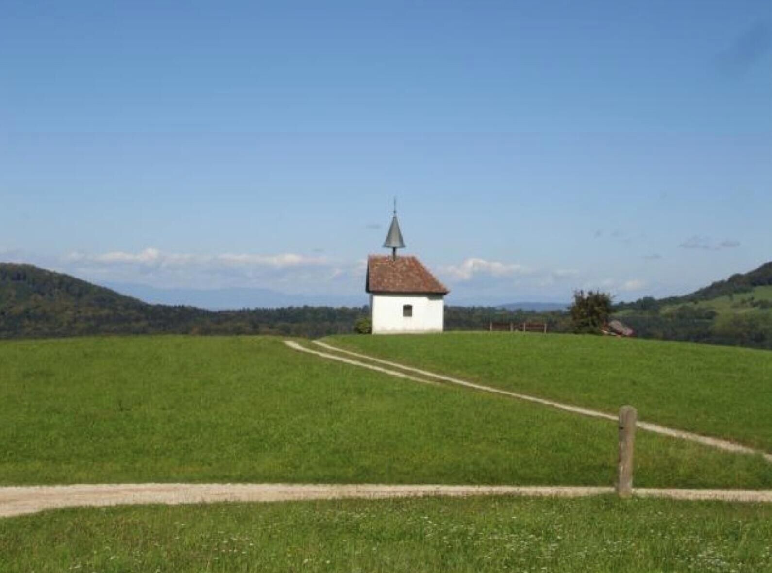 A small white building with a red roof and a pointed roof structure, surrounded by green fields and a clear sky.