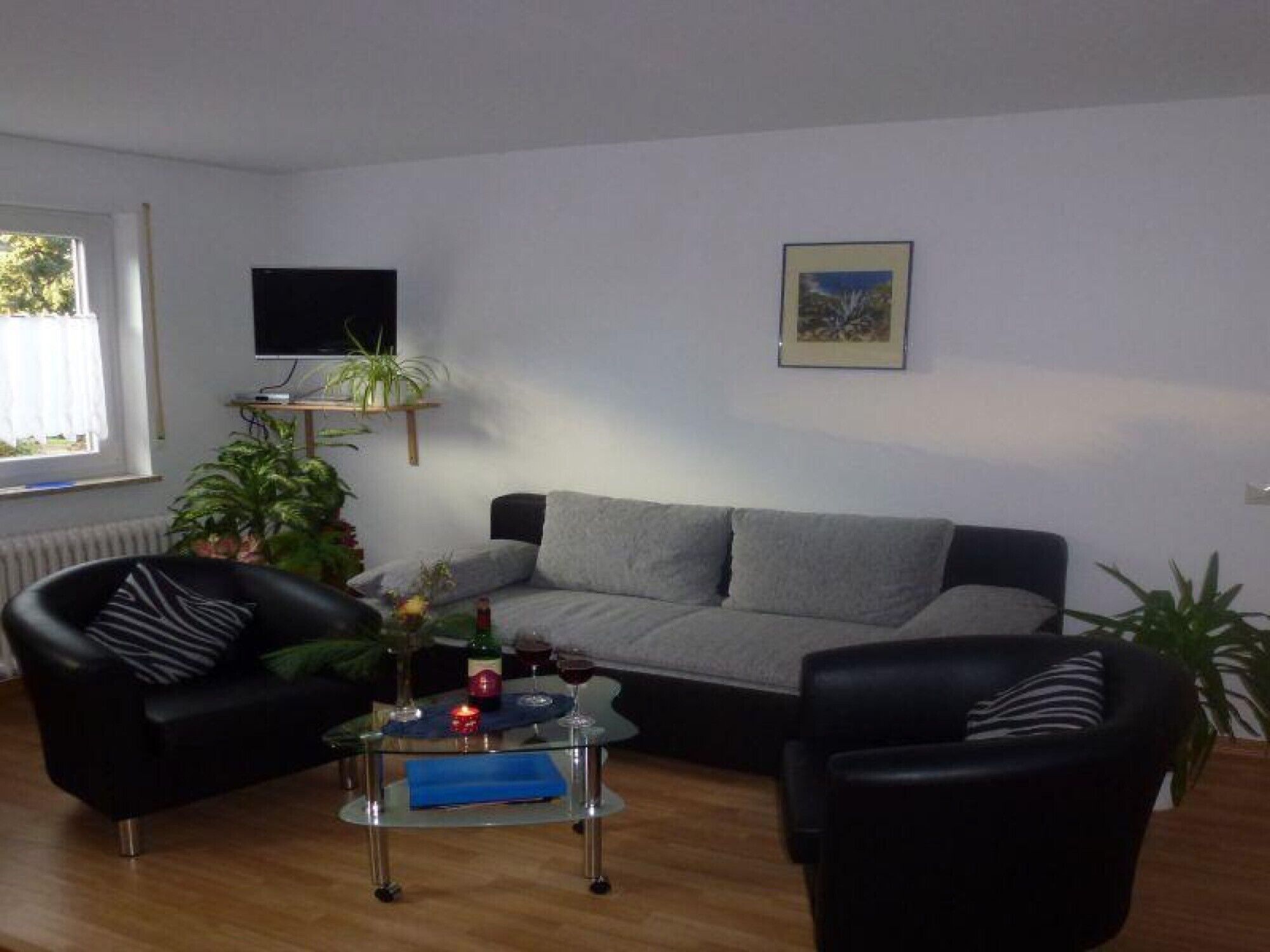 A living room with a grey sofa, two black armchairs, a glass coffee table, a TV on a stand, and a framed picture on the wall.