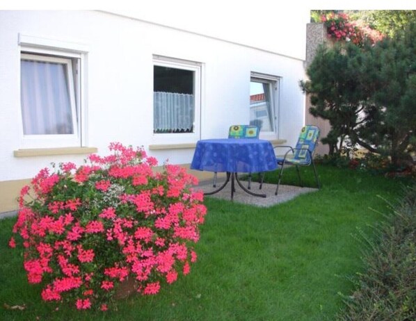 Un bâtiment blanc avec deux fenêtres, une petite table avec des chaises et un arbuste aux fleurs roses.