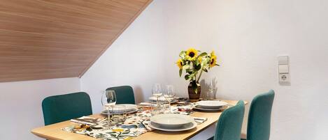 A dining area with a wooden ceiling, a table set for four with chairs, a vase of sunflowers, and a white wall with a power outlet.