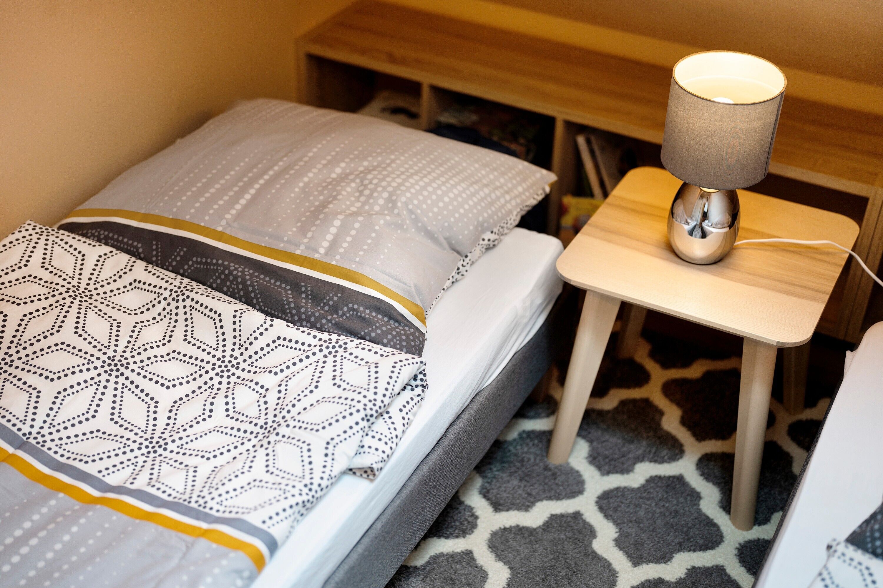 Een opgemaakt bed met beddengoed met een patroon, een nachtkastje met een lamp en een klein houten nachttafeltje.