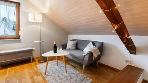 Acogedor dormitorio en ático con techo inclinado, un sofá gris, una pequeña mesa redonda con una botella y vasos, una lámpara de pie y un banco.