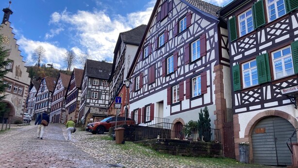 Una calle adoquinada flanqueada por edificios tradicionales con entramado de madera, una torre de iglesia y un letrero de un restaurante local.