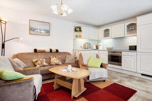 A living room with a sofa, armchairs, a coffee table, and a red rug. There is a kitchen with white cabinets and a tiled backsplash.