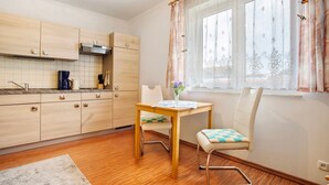Una cocina con armarios de madera, una mesa de comedor con sillas, un ventanal con cortinas y un pequeño jarrón con flores.