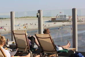 Uitzicht op het strand vanuit een kamer met twee houten stoelen, een raam en een tafel met een laptop.