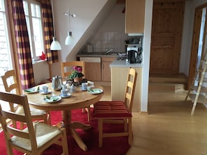 Een eetgedeelte met een ronde tafel, stoelen en een rood tapijt. In de achtergrond een keuken met houten kastjes en een koffieapparaat.