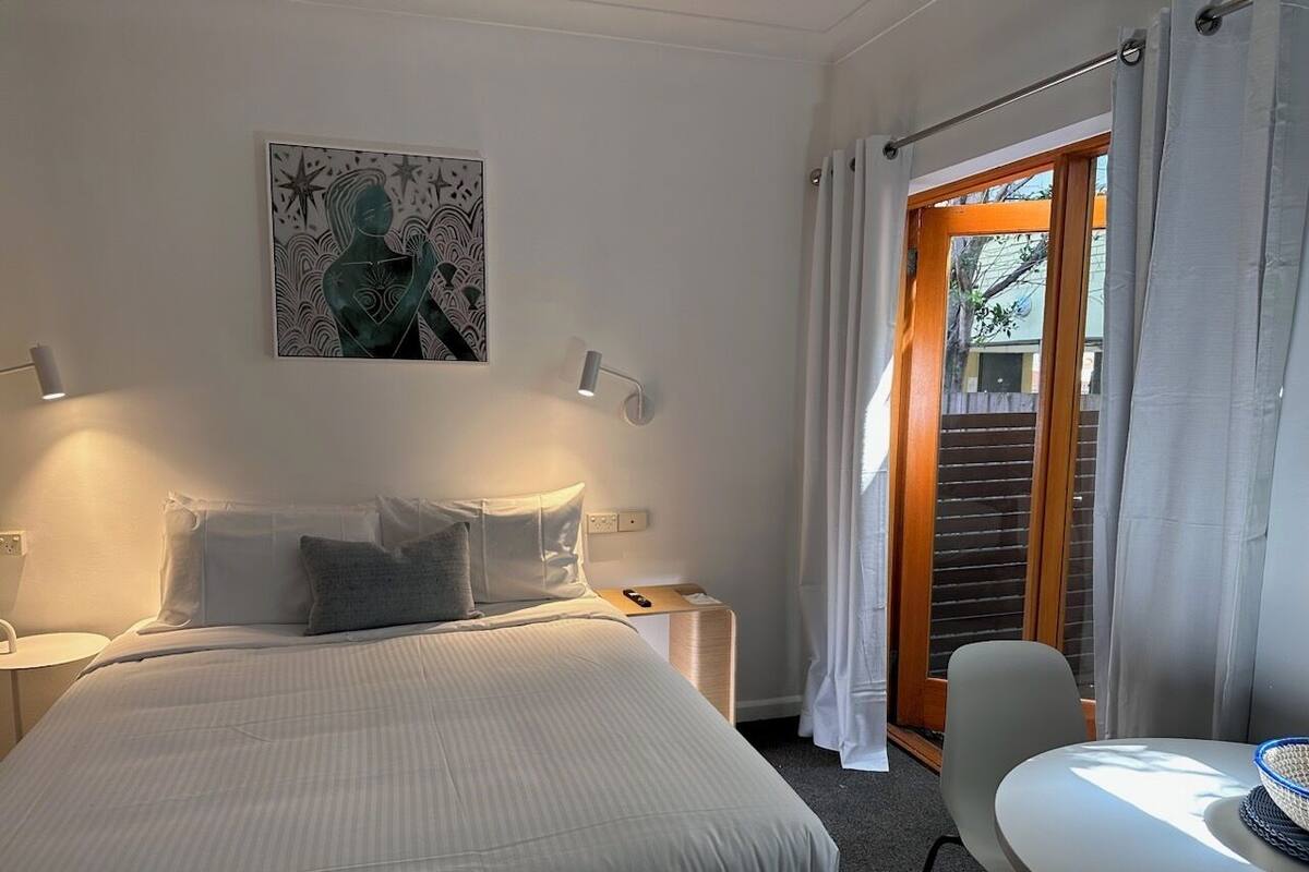 Habitación de hotel con cama, mesitas de noche, escritorio y una silla. Se ve una ventana con cortinas y vistas al exterior.