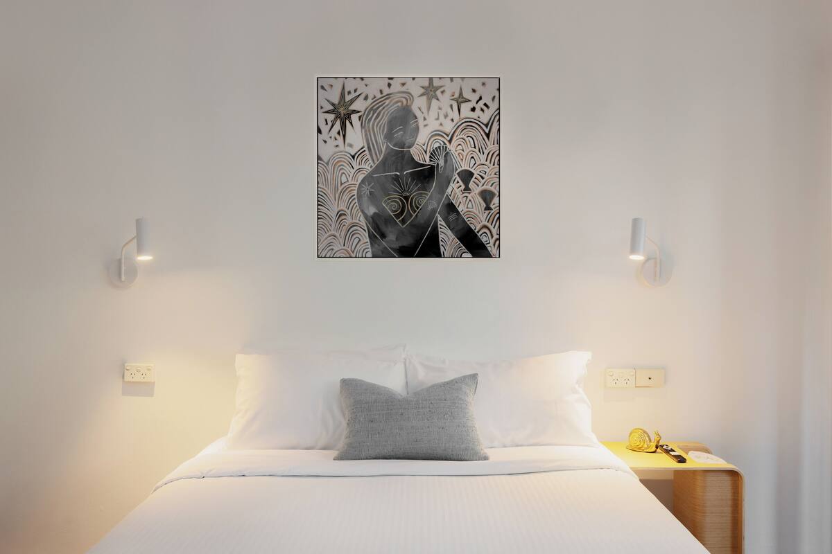 Una cama bien hecha con sábanas blancas y un cojín gris, una obra de arte colgada en la pared encima, y una mesita de noche con una lámpara y un teléfono.