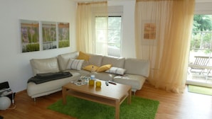 Una sala de estar con sofá, mesa de centro y alfombra verde. Hay cortinas en las ventanas y cuadros en la pared.