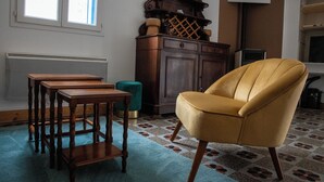 Een kamer met een gele fauteuil, houten tafels, een groene poef en een kamer met een patroon vloer.