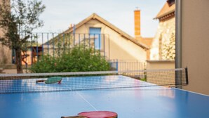 Een blauwe tafeltennistafel met een batje op de voorgrond, een net in het midden en een gebouw met een blauwe deur op de achtergrond.