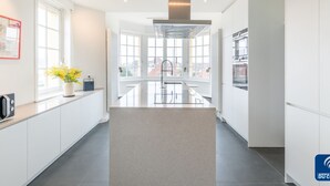 A modern kitchen with a central island, large windows, and a view of buildings outside. - Beautiful Normandy Villa in an Exclusive Location (De Haan)