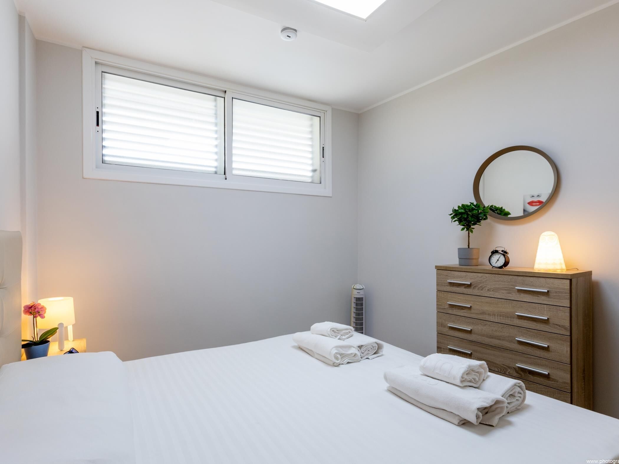 A bedroom with a bed, a dresser, a mirror, a window with blinds, and a bedside lamp.