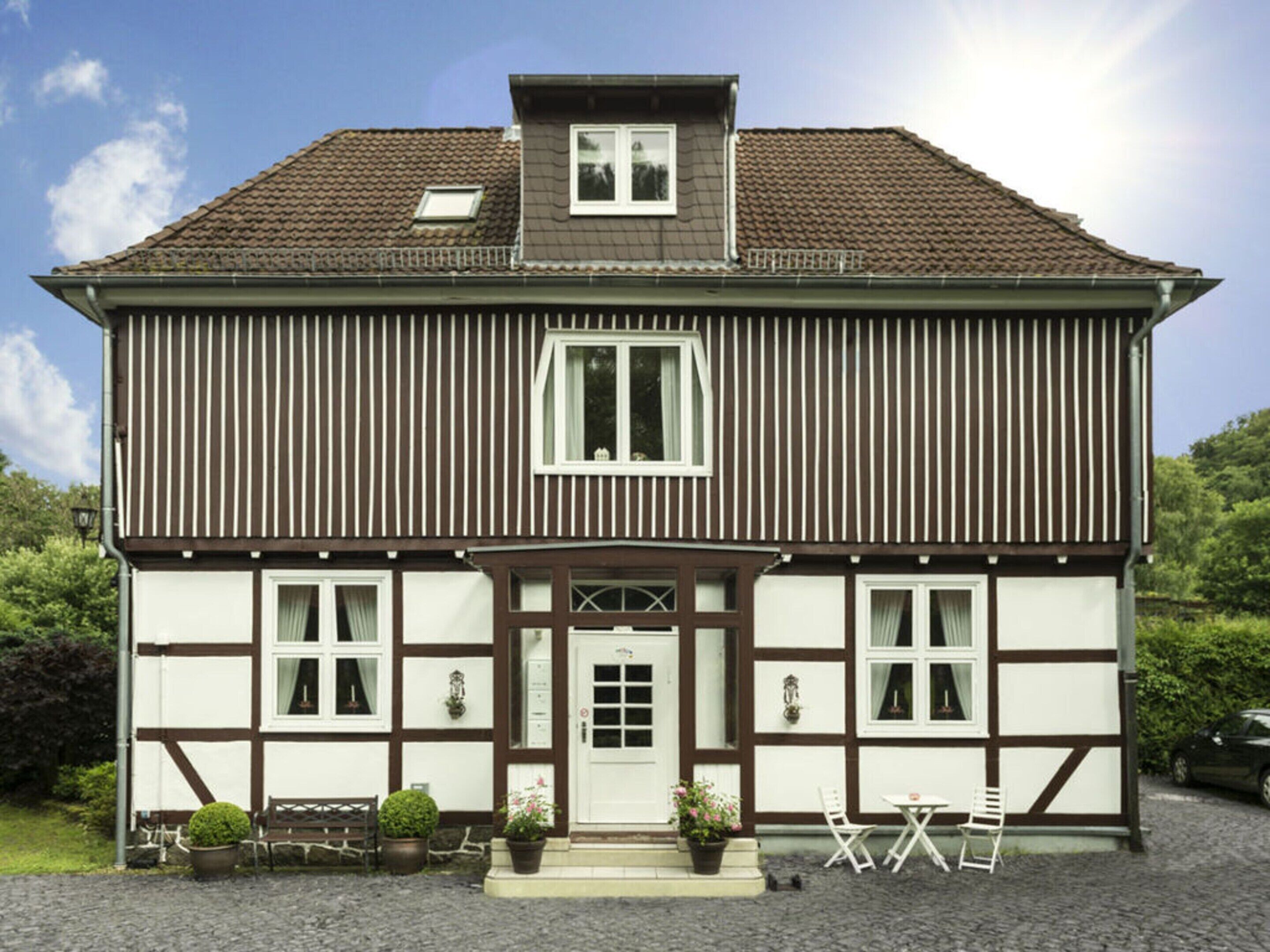 A traditional half-timbered house with a central entrance, a small porch with chairs, and potted plants.
