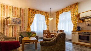 Een gezellige woonkamer met een bank, fauteuil en een eettafel. Er is een keuken met een fornuis en kasten.