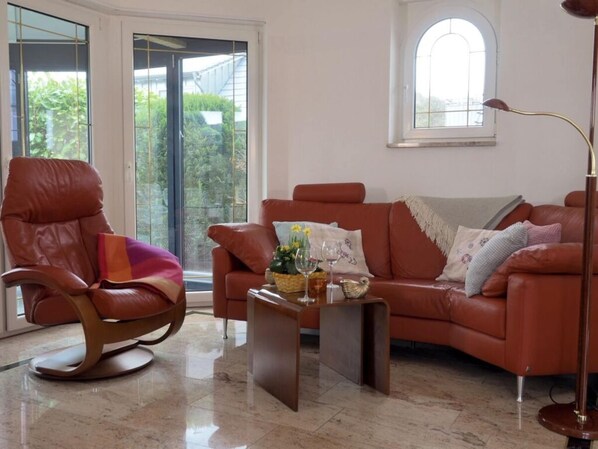 Ein Wohnzimmer mit Ledersessel, braunem Sofa und Couchtisch. Ein großes Fenster bietet Blick auf grüne Landschaft.