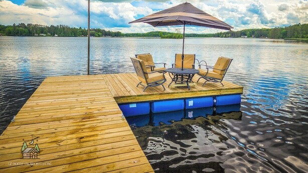 Un muelle de madera que se adentra en un lago, con una mesa y sillas bajo una pérgola.