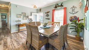 A dining area with a wooden table and wicker chairs, a kitchen in the background, and decorative wall art. - 1 Mi to Marina: Rockport Vacation Rental (Rockport)