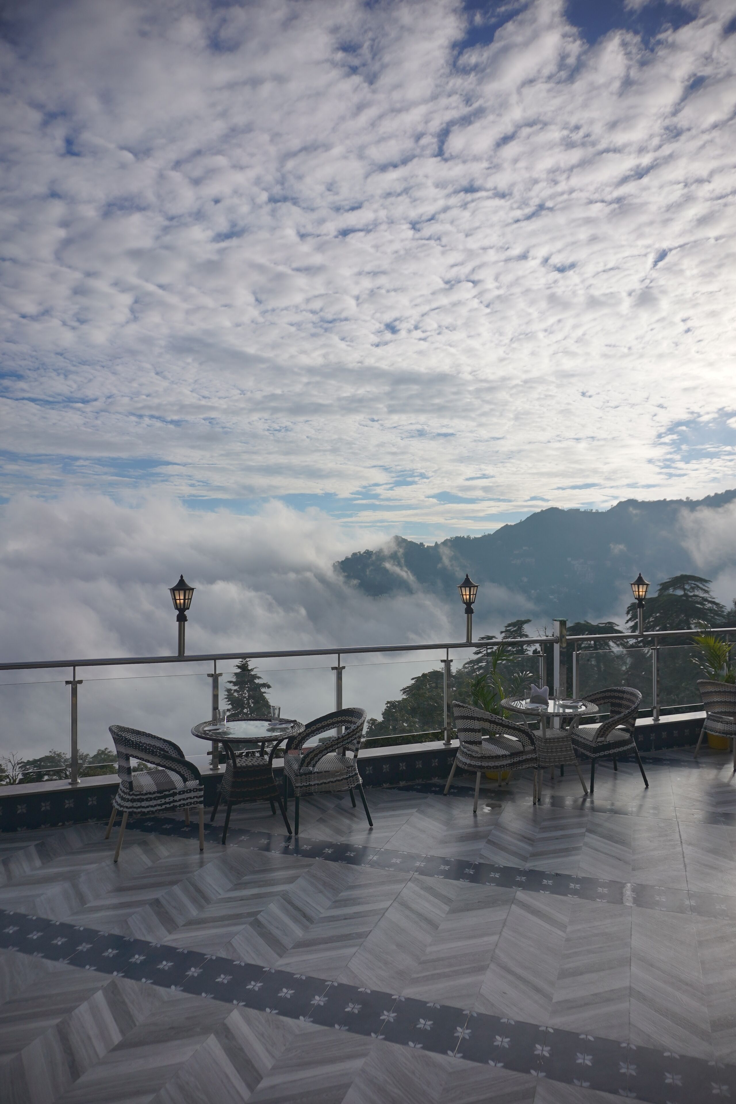 A terrace with wicker chairs and tables, overlooking a mountainous landscape with clouds below.