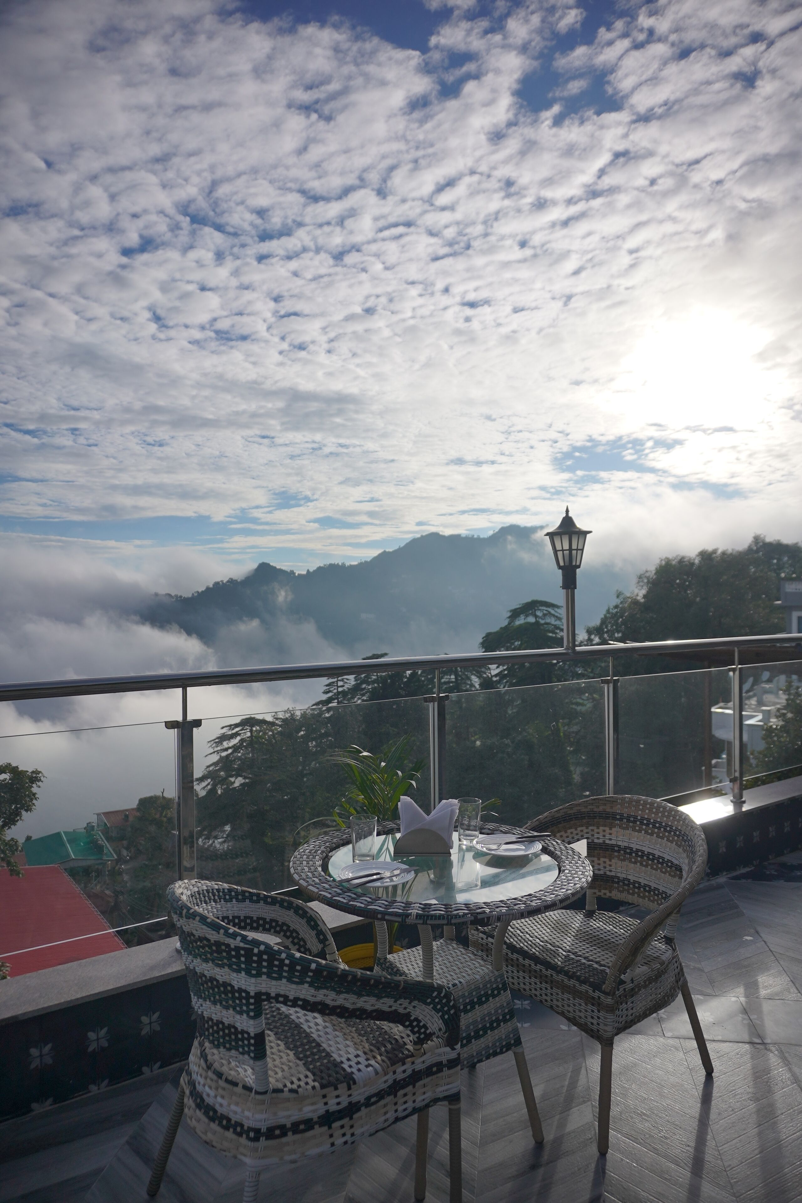 A balcony with wicker chairs and a glass table, overlooking a mountainous landscape with a view of the sea and clouds.