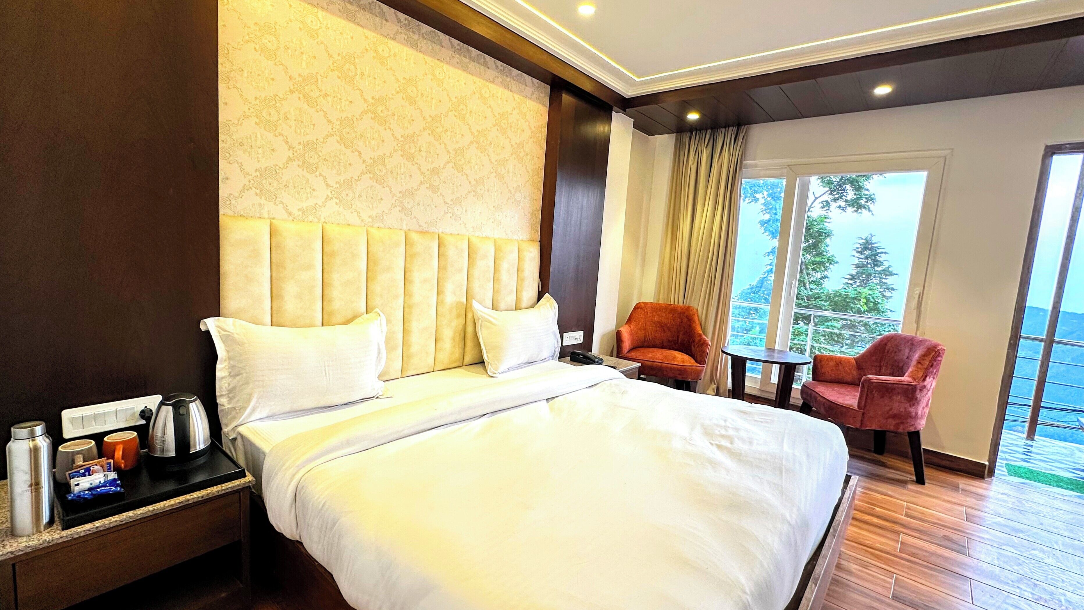 Habitación de hotel con una cama grande, una mesita de noche con tetera y tazas, una silla y vistas al exterior.