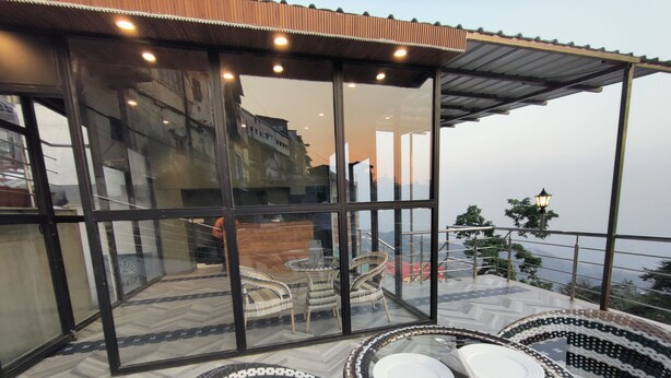 Un espace extérieur aménagé avec des meubles en osier, comprenant des chaises et une table, le tout protégé par des parois de verre, et offrant une vue sur le paysage urbain.