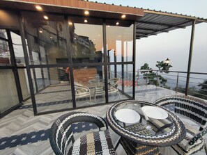 A glass-enclosed outdoor seating area with wicker chairs and a table, overlooking a cityscape. - Sangam by Stotrak (Dehradun)