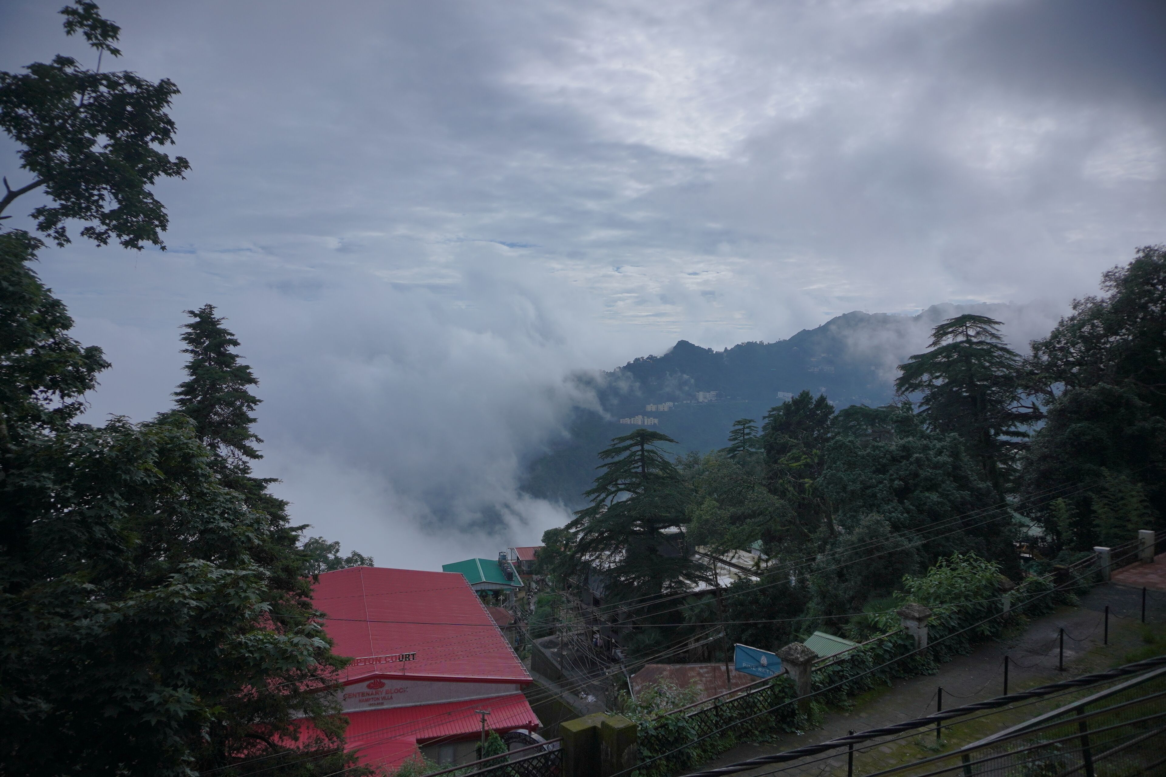 A mountainous landscape with a red-roofed building, dense greenery, and a cloudy sky.