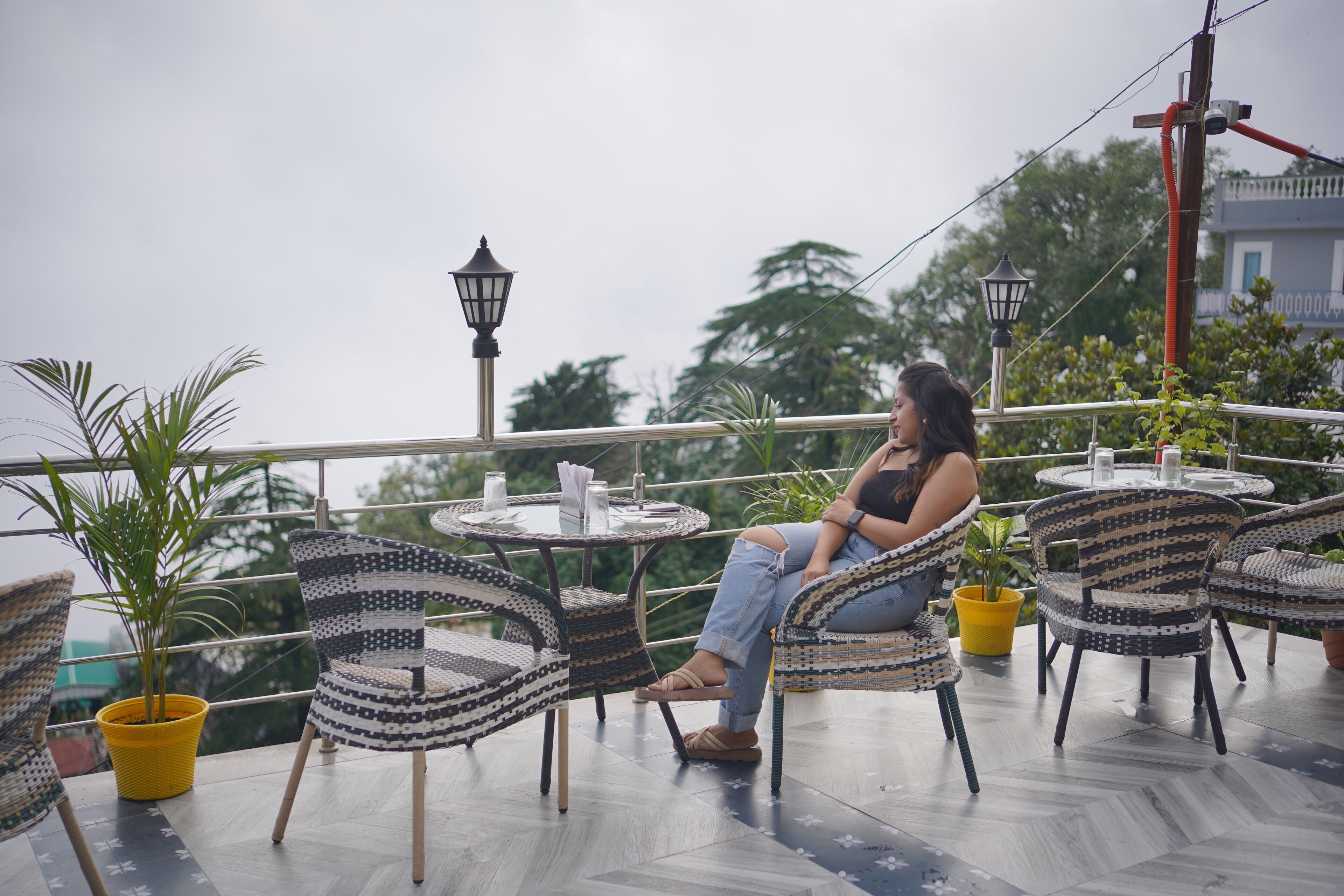 A woman sitting on a wicker chair on a balcony with a view of trees and a building in the background.