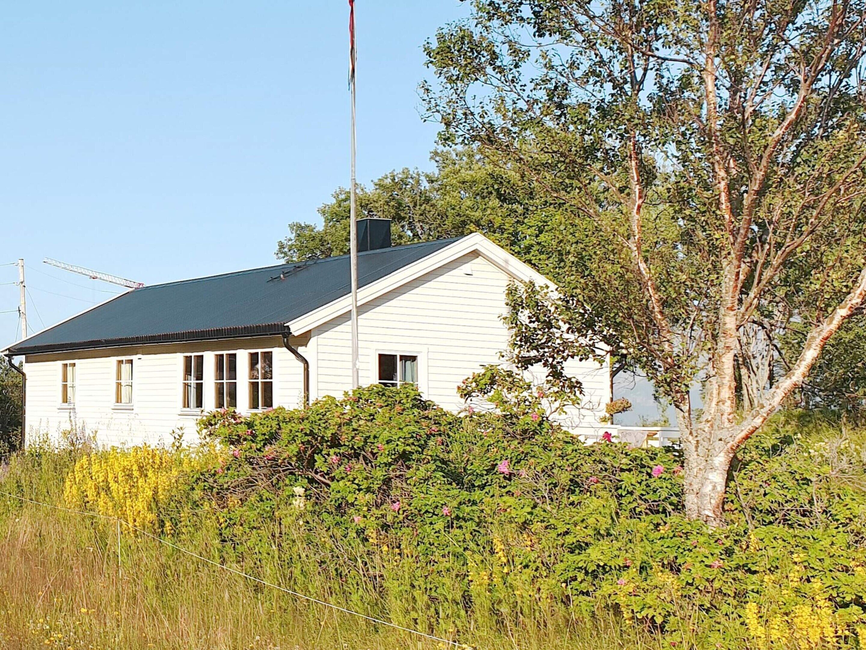 A white house with a dark roof, surrounded by dense greenery and a tall tree.