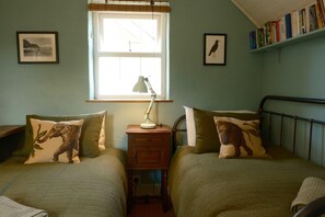 Ein Zimmer mit zwei Betten, jedes mit einem Kissen mit Elefantmuster, einem Nachttisch mit Lampe und einem Fenster mit Jalousien.