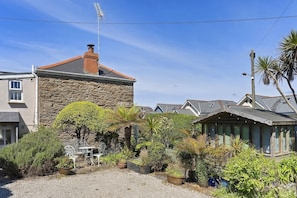 Uma casa de pedra com chaminé, rodeada por um jardim de cascalho com vasos de plantas e cadeiras de metal.