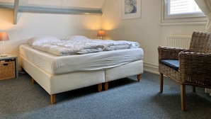 A bedroom with a bed, two bedside lamps, a wicker chair, a window with curtains, and a framed picture on the wall.