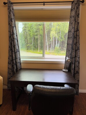 A desk with a chair in front, a window with blinds, and a view of a forest. - Cozy 1-bedroom apartment in charming North Pole with WiFi (North Pole)