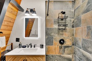 A bathroom with a white sink, mirror, and wooden vanity. It features a stone shower wall and a black towel rack.