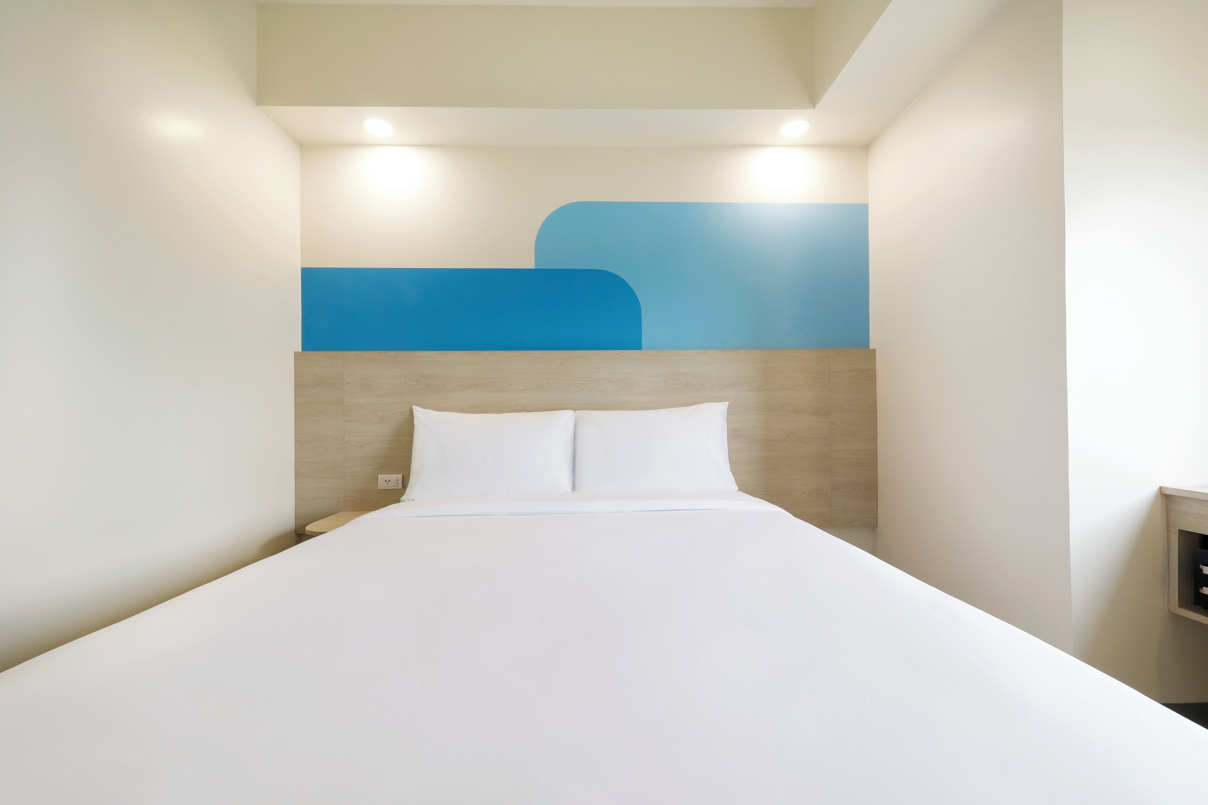 Un lit bien fait dans une chambre d’hôtel, avec une simple tête de lit et un luminaire fixé au mur.