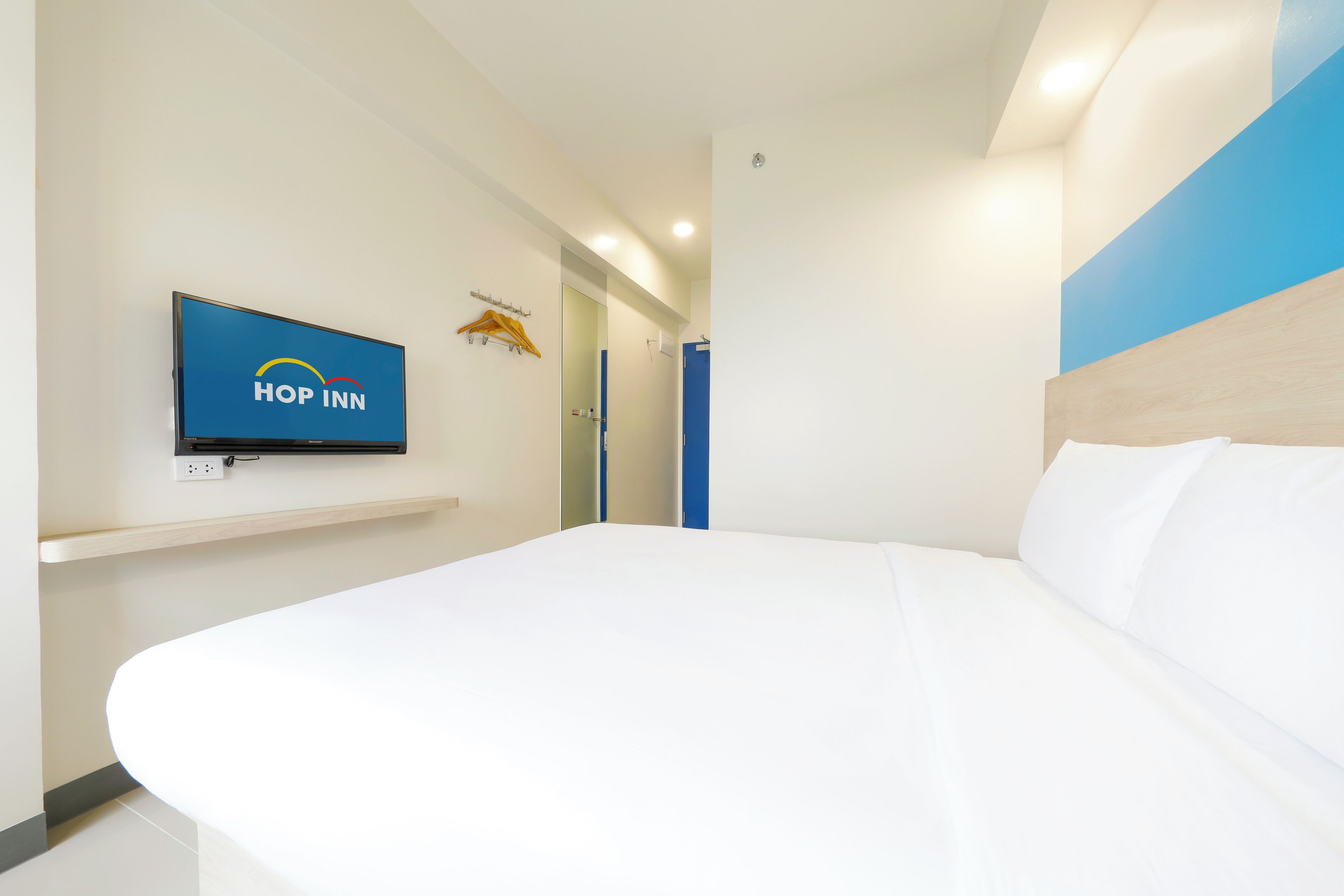 Una habitación de hotel moderna con una cama grande, un televisor montado que muestra el logotipo de 'Hop Inn' y una pared azul decorativa.