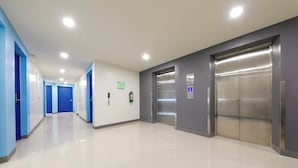 Un couloir moderne et bien éclairé, avec des portes bleues, un sol impeccable et un ascenseur.