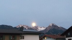 A mountain range with a visible moon in the sky, houses with sloped roofs, and snow-capped peaks. - Reindler Modern Retreat (Fischen im Allgaeu)