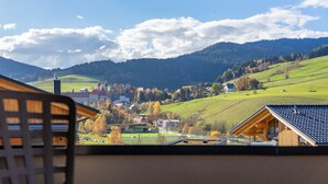 Vue depuis l’intérieur d’un bâtiment, donnant sur un village avec des constructions traditionnelles, une église et des collines ondoyantes.