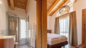 Une chambre moderne avec un plafond en bois, une grande fenêtre avec des rideaux, un lit et une salle de bain dotée d’une cabine de douche en verre.